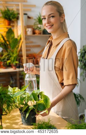 Vertical Portait Of Smiling Woman Florist In Apron Spraying Water On Houseplants In Pots By Sprayer,
