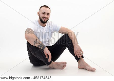 Young Smiling Bearded Man With Short Dark Hair In White T-shirt, Black Jeans, Sitting, Bending Knees