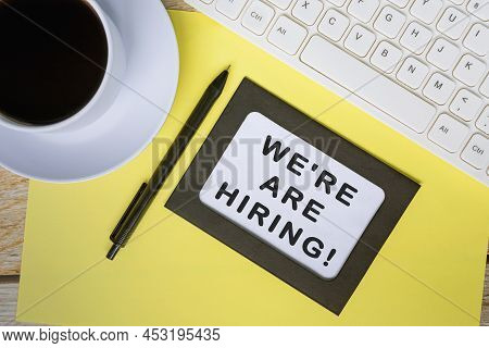 We're Are Hiring Text On White Note With Keyboard, Pen And Coffee Cup On Yellow Background.