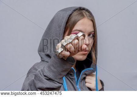 A Teenage Girl Strikes With Brass Knuckles On A Light Background, Wearing A Hoodie With A Hood. Conc