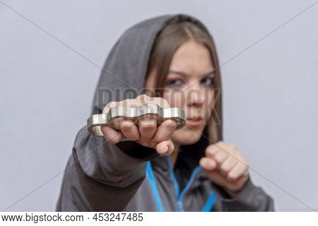 A teenage girl strikes with brass knuckles on a light background, wearing a hoodie with a hood. Conc