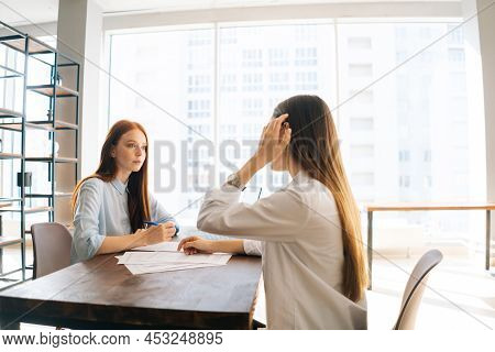 Rear View Of Two Female Office Workers Sitting At Desk With Paper Documents Looking At Each Other Di