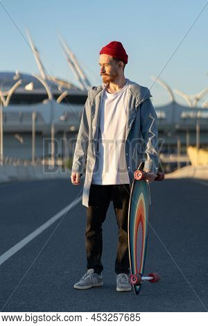 Casual Man With Longboard On Sunset Over Modern City Building. Hipster Skater Holding Skateboard Wea