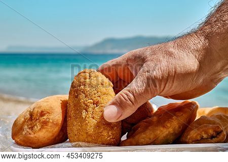 Typical Sicilian Street Food On Seaside Background. Hand Of Man Taking Arancina (deep Fried Rice Bal