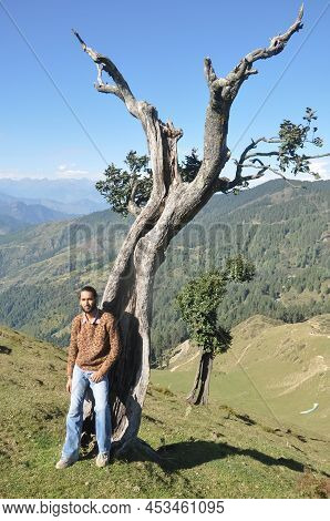 Full Length Of A Stylish Indian Young Man Looking At Camera While Posing With Leaning Against Tree I