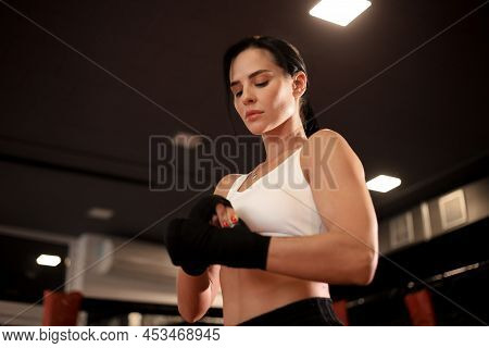 Young Woman Wraps Her Fists In Black Bandages For Kickboxing Gloves Against A Black Boxing Hall