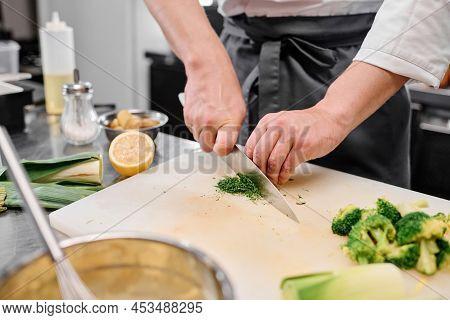 Close-up Of Chef Using Kitchen Knife To Cut Broccoli In Little Pieces For Dish On Cutting Board At T