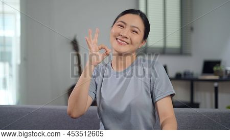 Smiling Confident Asian Woman Showing Ok Sign By Hand Sitting On Sofa In Living Room At Home Looking
