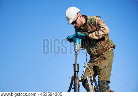 Male Worker Building Pile Foundation For Wooden Frame House. Man Builder In White Safety Helmet Dril