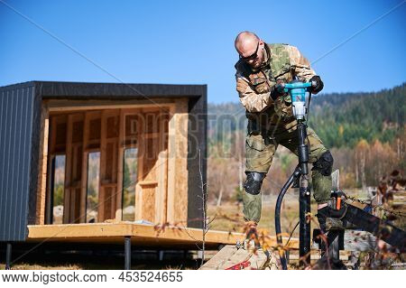 Male Workers Building Pile Foundation For Wooden Frame House. Men Builders Drilling Piles Into The G