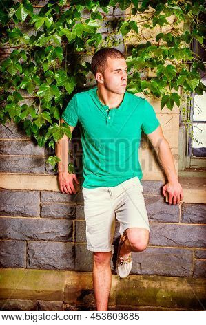 Young Man Waiting. Wearing A Green Short Sleeve Henley Shirt, Light Yellow Shorts, Bending A Leg, A 