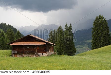 Wooden Hut On The Slope Of The Forest-covered Mountains In The Swiss Alps Under Dramatic Clouds On R