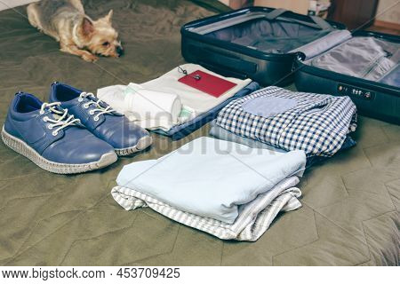 Stacked Clothes, A Passport, A Pair Of Shoes, And Personal Hygiene Items Next To An Open Suitcase