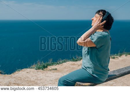 Portrait Adult Woman Listens To Music In Headphones In Summer, Spring Against The Backdrop Of Mounta
