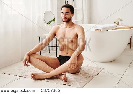 Young Shirtless Man Sitting On Carpet In Lotus Position And Breathing Deeply During Meditation In Ba