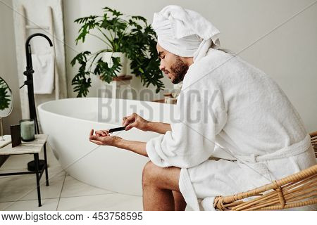 Young Handsome Man In Bathrobe Doing Manicure For Himself With Nail File While Sitting On Chair In B