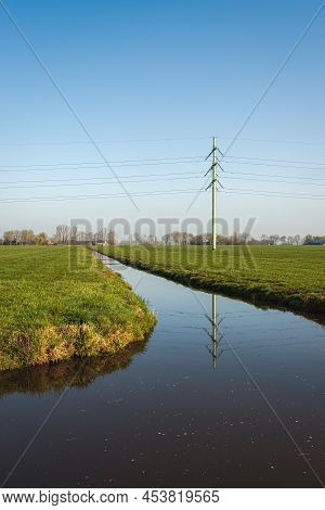 Dutch Polder Landscape With One High Voltage Pylon With High Voltage Cables Against A Blue Sky. The 