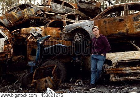 Ukrainian Man Stands Against The Background Of Broken And Burnt Cars Affected By Russian Missiles. D