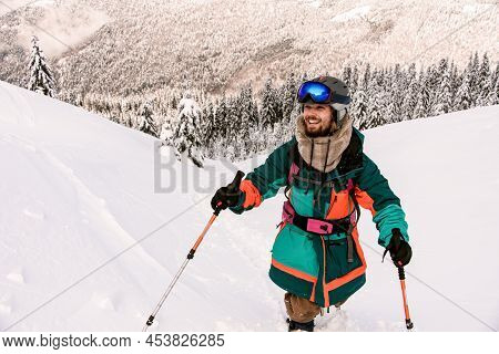 Happy Smiling Skier In Bright Jacket Is Climbing The Hill Using Skitour Equipment.