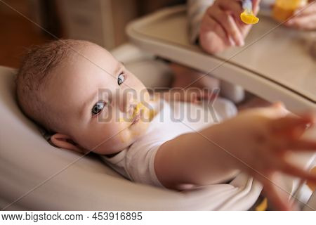 Mother Feeding Her Baby Boy Using Spoon, Introducing First Solid Food Meal, Baby Sitting In High Cha