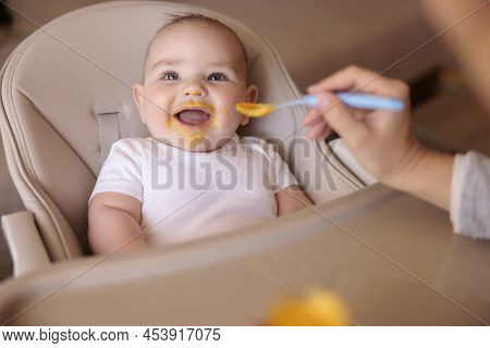 Mother Feeding Her Cheerful Baby Boy While Introducing First Solid Food Meal, Baby All Messy And Sta