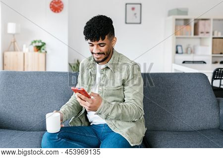 Young Hispanic Latino Man Using Mobile Phone While Sitting On A Sofa At Home - Cheerful Millennial V