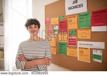 Happy Young Woman Student Standing In Front Of Employment Noticeboard.