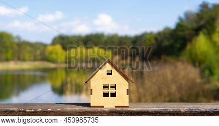 Miniature Toy Wooden Cabin Model Standing Against A Beautiful Lake Landscape Background. Buying A La