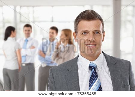 Middle-aged businessman in front, business team talking in background.