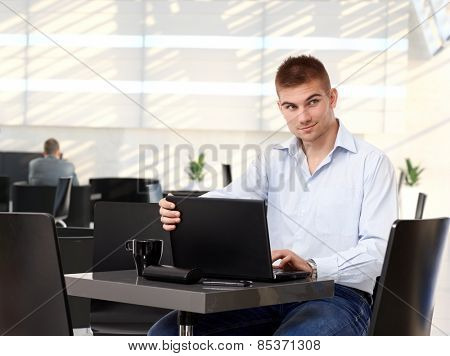 Young casual rookie businessman working on laptop computer at cafe, smirking, copyspace, sitting at table.