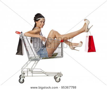 Happy young beautiful brunette woman sits in an empty shopping cart with a red bagisolated on white background