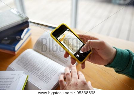 people, education, technology and exam concept - close up of student hands with smartphone taking picture of books page and making cheat sheet in school library