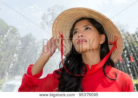 Vietnamese woman in a traditional red outfit and conical hat standing in front of a fountain