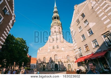 Riga Latvia - July 1 2016: Bottom View Of St. Peter Church Main Facade With Fleche Full Height Between Two Old Buildings Blue Clear Sky Background. Walking People In Frame.