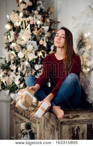 Woman with a Christmas Gift sitting on acncient commode decorated for xmas. Fir tree at background.