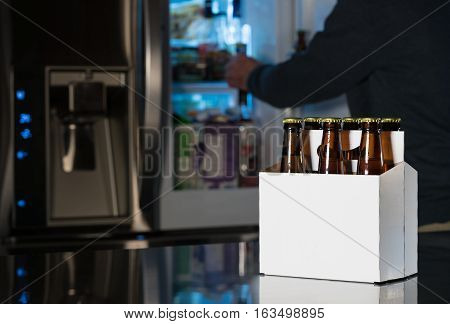 Six pack of brown beer bottles in plain white cardboard carrier with copy space on stainless steel kitchen or bar counter. Open fridge or refrigerator out of focus in rear.