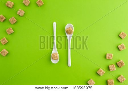 Brown sugar cubes on greenery background with two white teaspoons in the middle. Top view. Copy space for text. Diet unhealty sweet addiction concept