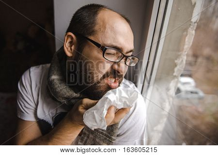 Young man blowing his nose while sitting at the window
