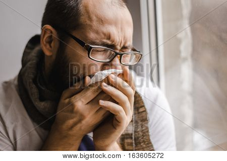 Young man blowing his nose while sitting at the window