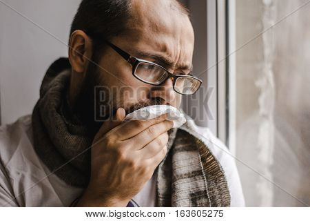 Young man blowing his nose while sitting at the window