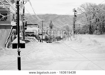 A quiet, early morning street in Myoko. Snow-covered buildings, cars and roads from the overnight snowfall. Distant mountains show the extent of the snowfall. Black and white image.