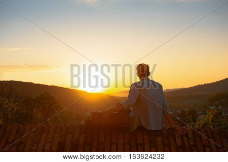The boy who seek a new beginning. Admire the sunset sitting on a fence made of brick with luggage and ready to go.