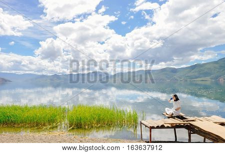 Single young Chinese woman in white long skirt sits on wasted platform on bank of Lugu lake, listening to music, back to camera, at Daluoshui village, Lijiang, China