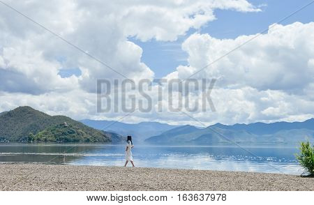 Single young Chinese woman in white long skirt sits walks on beach along the bank of Lugu lake, at Daluoshui village, Lijiang, China