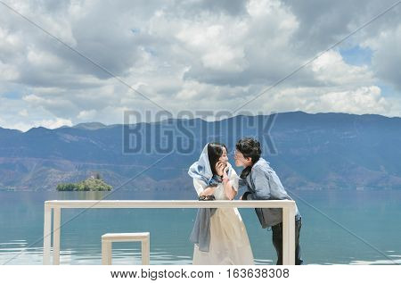 Happy young Chinese couple leans on white long table looking at each other, by the bank of Lugu lake, at Daluoshui village, Lijiang, China
