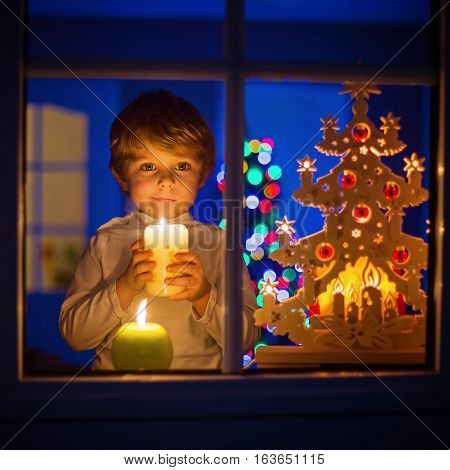 Beautiful little kid boy, standing by window at Christmas time and holding candle. Child waitng for Santa. With colorful lights from Christmas tree on background.