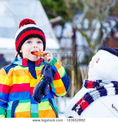 Adorable little kid boy making a snowman and eating carrot. child playing and having fun with snow on cold day. Active outdoors leisure with kids in winter.