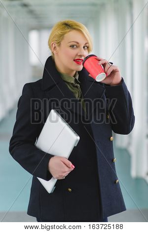 Portrait of blond businesswoman holding coffee and laptop in lobby.