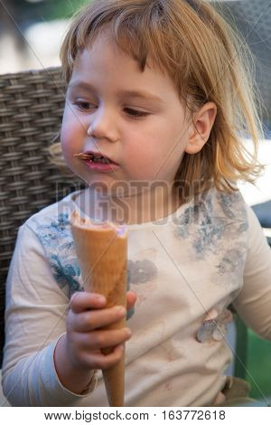 portrait of two years old blonde cute child with white shirt sitting and biting strawberry ice cream cone at urban park