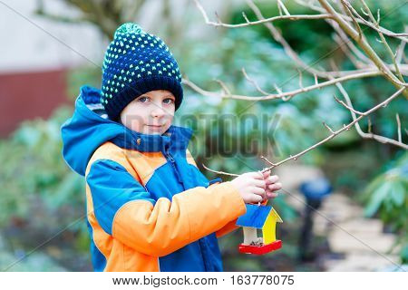 Little kid boy feeding birds in winter. Cute happy preschool child hanging colorful selfmade bird house on tree on frosty cold day. Preschooler in colorful wam clothes. Nature, empathy with animals.
